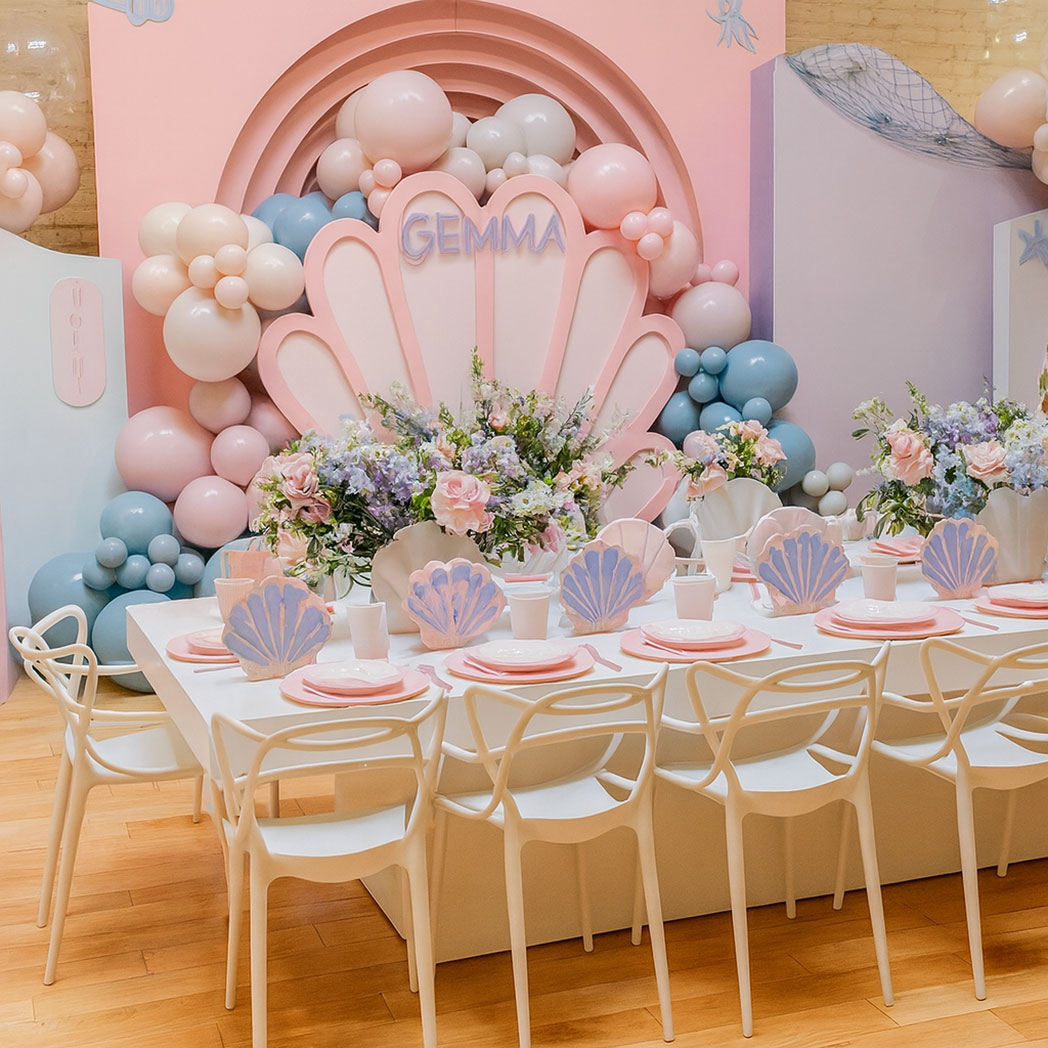 Modern acrylic kids dining table styled for a mermaid theme birthday party with pastel balloons, seashell plates, and white chairs