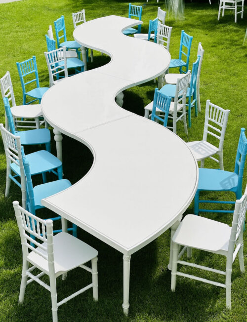 Kids serpentine event table with glossy white acrylic top and polished stainless steel frame surrounded by white and blue Chiavari chairs on green grass