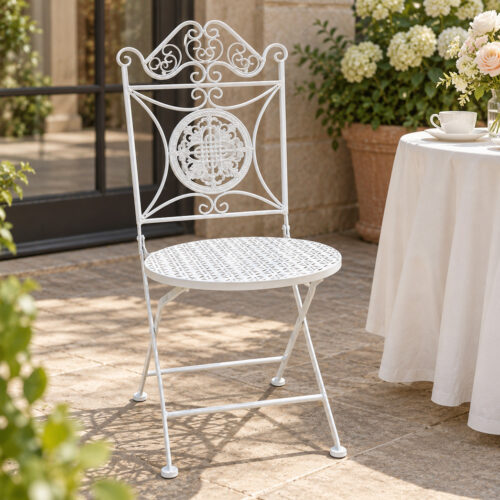 White Florentine scroll back wrought iron folding chair with ornate floral medallion and perforated round seat displayed on a sunlit garden patio beside a floral café table.